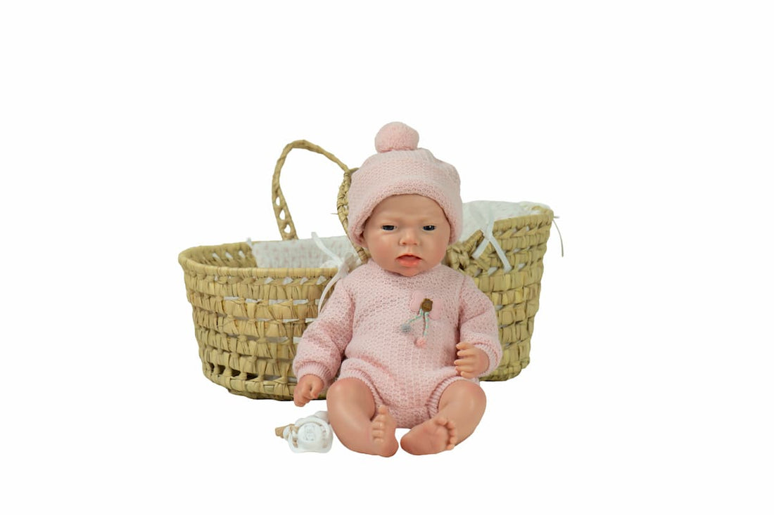 Bebé Real Reborn Arlet de Arias sentado junto a su capazo de mimbre, con vestido rosa y gorro blanco