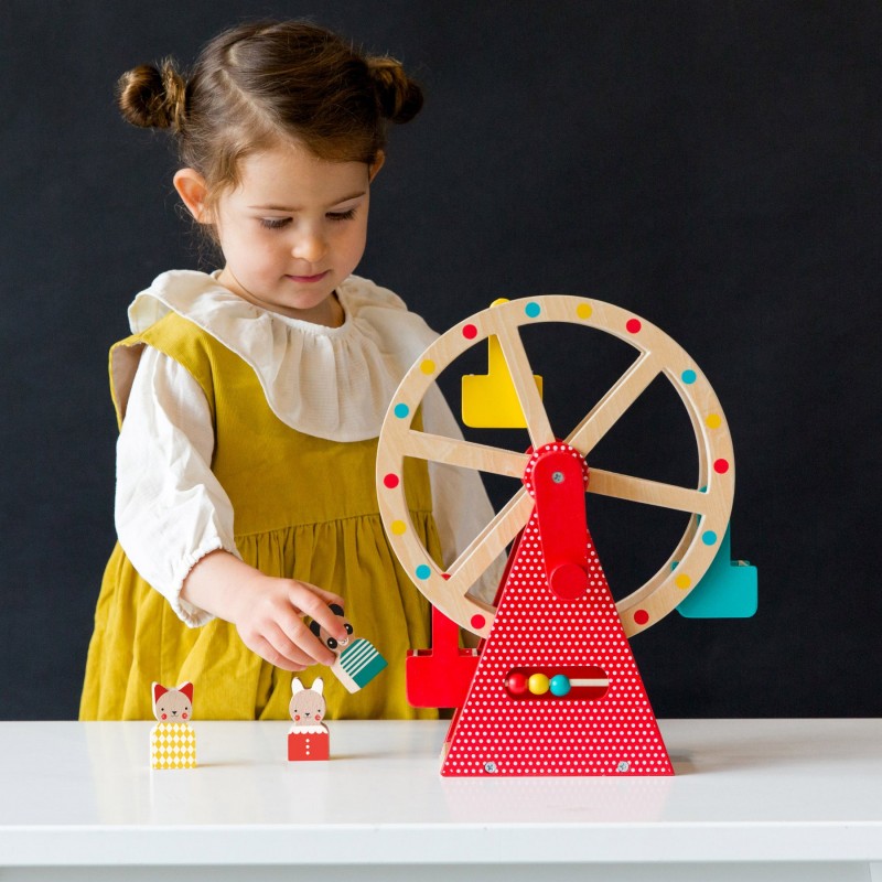 niña con vestido amarillo: Niña jugando con la gran noria de madera Petit Collage y sosteniendo una figura de panda junto a otros animales sobre la mesa.