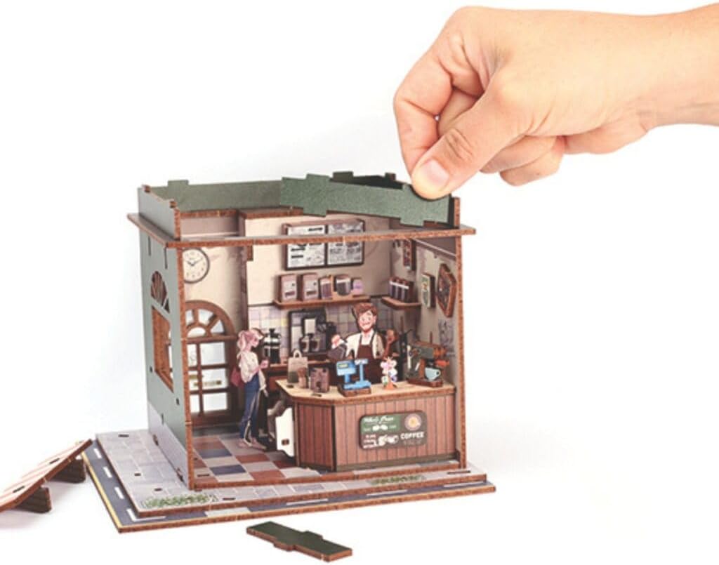 Montaje de la maqueta de madera 3D Coffee Shop de Graine Creative, con una mano colocando una pieza sobre la cafetería.