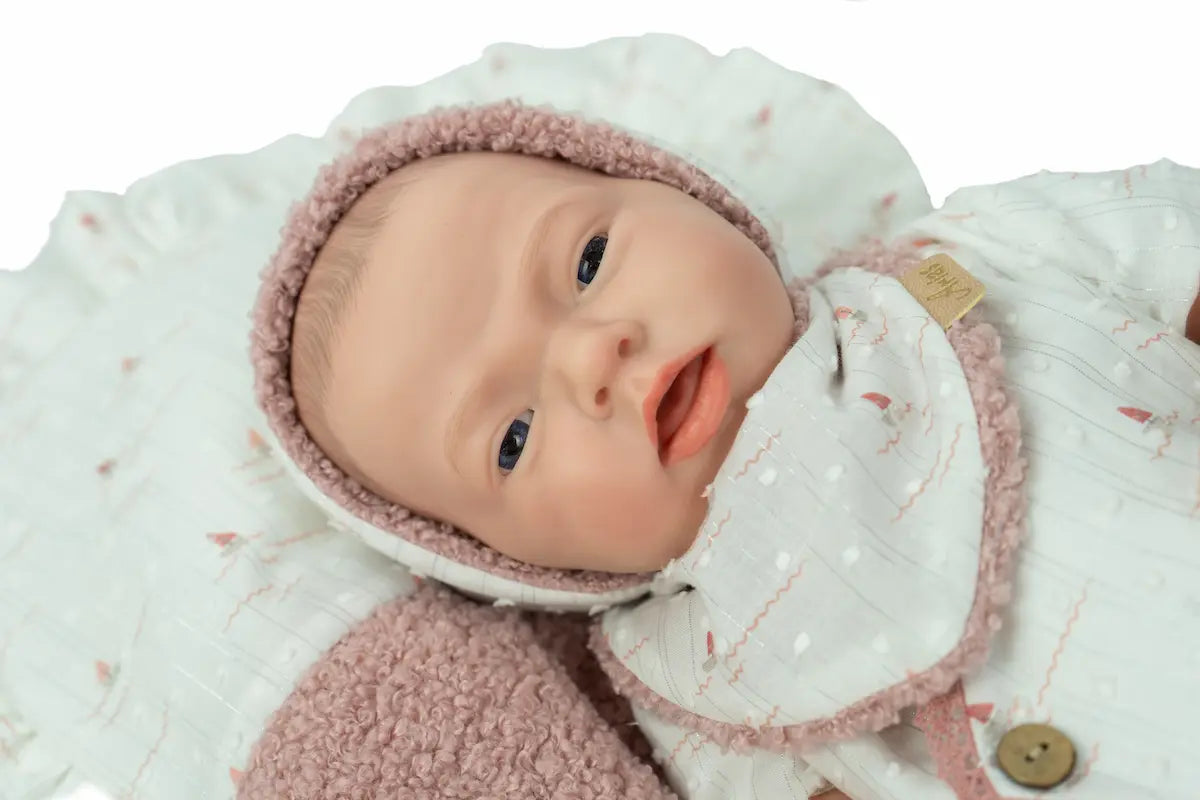 Reborn Aitana de Arias 98204 tumbada boca arriba con gorrito blanco sobre una manta clara.